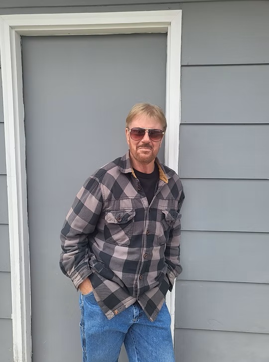 A man in sunglasses and a plaid shirt stands with hands in pockets in front of a gray door and wall.